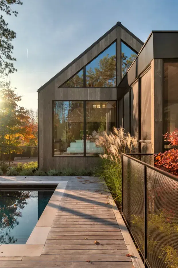 Exterior architectural photograph of a contemporary dark-clad house with a prominent triangular gable and expansive black-framed glazing. A timber deck and walkway border a reflecting swimming pool, with planted grasses and shrubs integrated around the building. Warm late-afternoon light highlights the textured dark facade, timber decking, and landscaped outdoor setting.