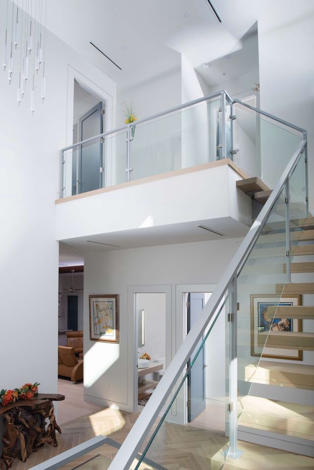 Bright contemporary interior view centered on a stairway rising beside a mezzanine or upper landing. The stairs have pale timber treads, clear glass balustrades, and slim metal handrails. White walls and ceilings, concealed linear lighting, and a light timber-edged mezzanine create an airy, minimalist composition. Living and dining areas are visible below and through doorways, with artwork and a sculptural timber side table adding warmth.