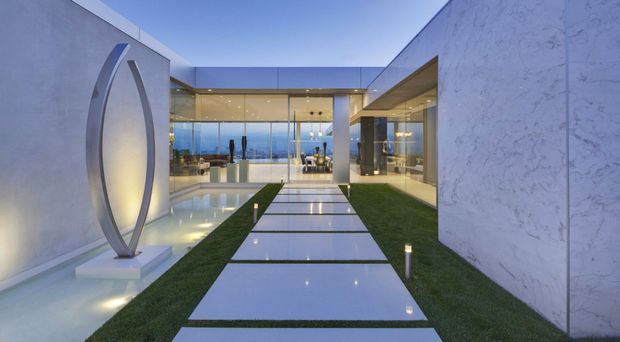 A twilight view through a highly contemporary courtyard toward a glass-fronted residence. Large pale stone or concrete walls frame a landscaped grass strip and a sequence of large rectangular stepping slabs, with slim illuminated bollard lights. A tall abstract oval metal sculpture stands in a shallow reflecting pool at left. Floor-to-ceiling glazing reveals a refined interior, while the distant blue horizon is visible beyond.