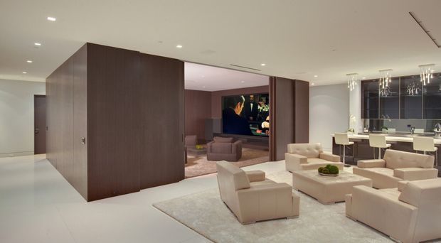 A spacious open-plan contemporary interior combining a living room, dining area and kitchen. The room features a large dark timber-clad central volume enclosing a media room, pale upholstered lounge furniture arranged around a low coffee table, a light neutral rug, recessed ceiling lighting, and a sophisticated kitchen with a dark splashback and island seating. The palette is predominantly warm neutral with dark timber and charcoal accents.