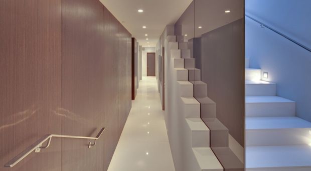 A narrow, minimalist interior hallway with glossy light flooring, warm wood wall panelling and a continuous handrail on the left. On the right, a sculptural stepped stair or shelving installation runs alongside a large reflective glazed surface, with a softly illuminated stairwell visible beyond.