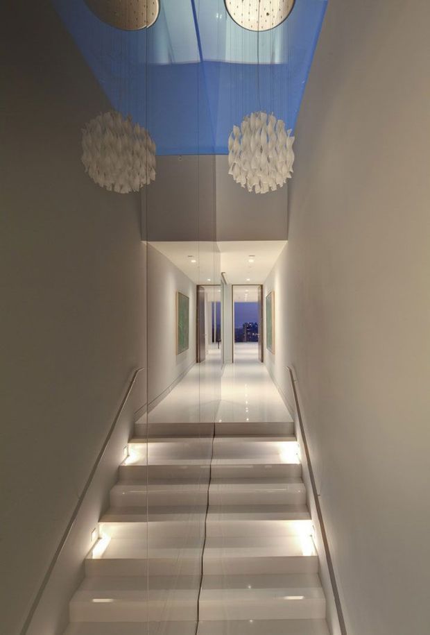 A narrow, refined interior stairway leading up to a long hallway. The glossy white stairs are illuminated beneath each tread, with slim handrails along the walls and mirrored or highly reflective wall panels. At the upper landing, recessed ceiling lights and framed artwork line the corridor, while a blue translucent ceiling feature and sculptural pendant lights create a dramatic focal point.