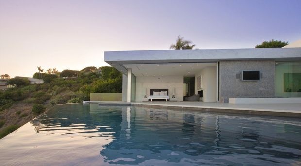 Exterior view of a sleek, single-storey modern residence beside an infinity-edge swimming pool. The home has a broad flat roof with deep overhangs, minimal rectangular forms, pale textured exterior walls, recessed ceiling downlights, and large open glazing or sliding doors connecting the living area to the pool terrace. The setting appears elevated with landscaped hillside views and warm dusk light.