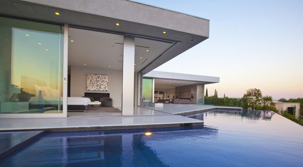 Exterior architectural view of a sleek, flat-roofed modern residence opening onto a large infinity-edge pool. The composition features expansive floor-to-ceiling glazing, projecting cantilevered roof slabs, crisp white and grey surfaces, recessed downlights, and a covered outdoor entertaining pavilion. A living area is visible through the glazing, with landscaped hedging and distant suburban surroundings beyond.