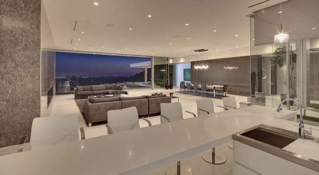 Open-plan luxury kitchen, dining, and living area with a long white island and integrated sink in the foreground, white bar stools, a large sectional seating arrangement, dining table, extensive glazing, and a nighttime city view. The space has a polished contemporary fit-out with recessed ceiling lights, dark textured feature walls, and reflective glass partitions.