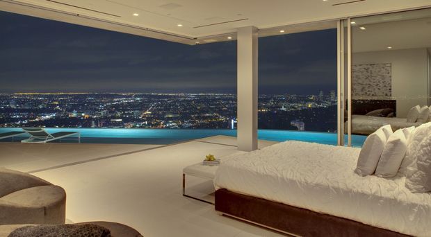 A luxurious contemporary bedroom with a large white bed, pale upholstered seating, and expansive floor-to-ceiling glazing overlooking a city and illuminated swimming pool at night. The room has a calm, refined palette of white, cream, beige, and dark brown, with recessed ceiling lighting and a mirrored or reflective glazed wall expanding the view.