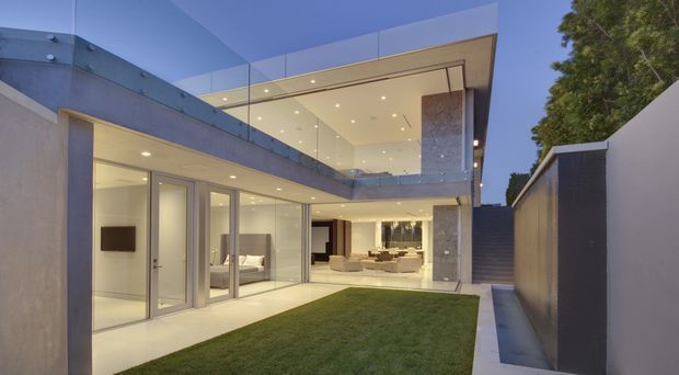 Twilight exterior view of a contemporary multi-level residence arranged around a compact lawn courtyard. The house features extensive floor-to-ceiling glazing, pale rendered walls, exposed concrete or stone-textured columns, a projecting upper-level volume with a glass balustrade, recessed downlights, and a narrow paved walkway along the boundary wall. Bright, minimalist interiors are visible through the glazing.