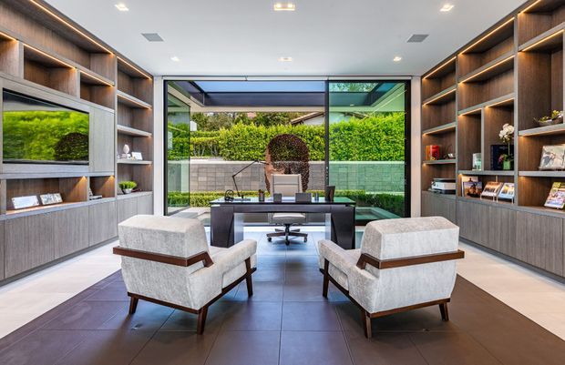 A refined home office or study with a central desk facing full-height glazing and a landscaped courtyard beyond. Two upholstered lounge chairs sit in the foreground, while extensive dark timber built-in shelving and cabinetry line both side walls, incorporating books, decor, and display niches. The room has a clean contemporary fit-out, recessed ceiling lighting, concealed warm LED shelf lighting, and large-format dark floor tiles.