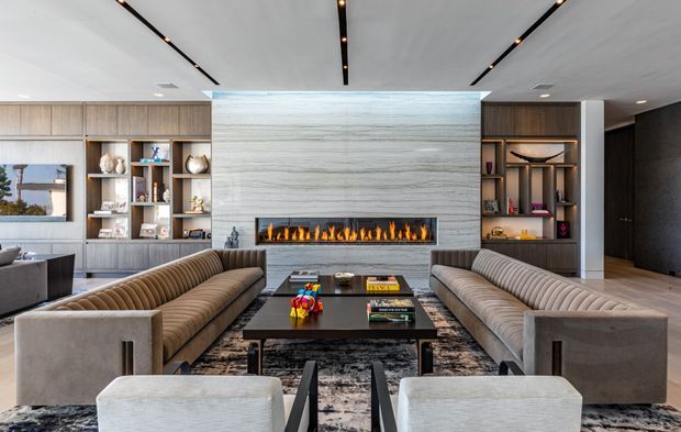 A refined contemporary living room centered on a long linear gas fireplace set into a large pale stone feature wall. Two matching ribbed sofas face each other across a dark rectangular coffee table and patterned rug, with built-in timber display cabinetry on both sides. Slim recessed linear lighting runs across the ceiling, and a dark doorway or passage is visible at right.