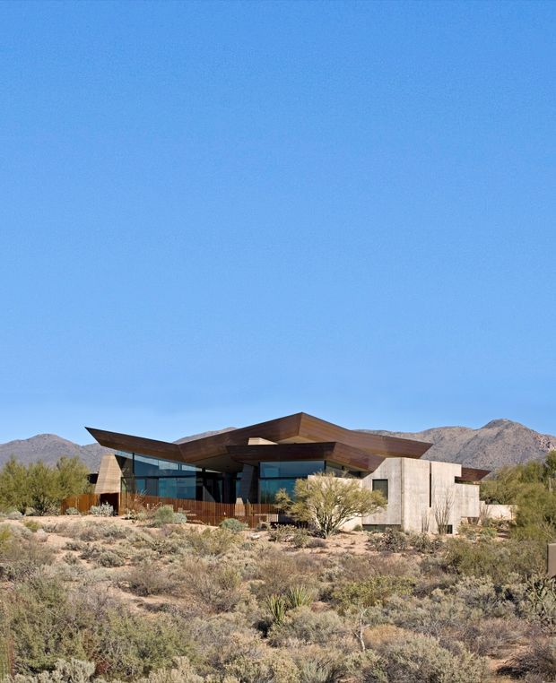 Low, angular modern house set in a dry desert landscape, with broad projecting roof planes, extensive dark glazing, warm timber cladding, and pale concrete walls. Sparse xeriscaped planting and rugged mountains form the surrounding setting beneath a clear blue sky.