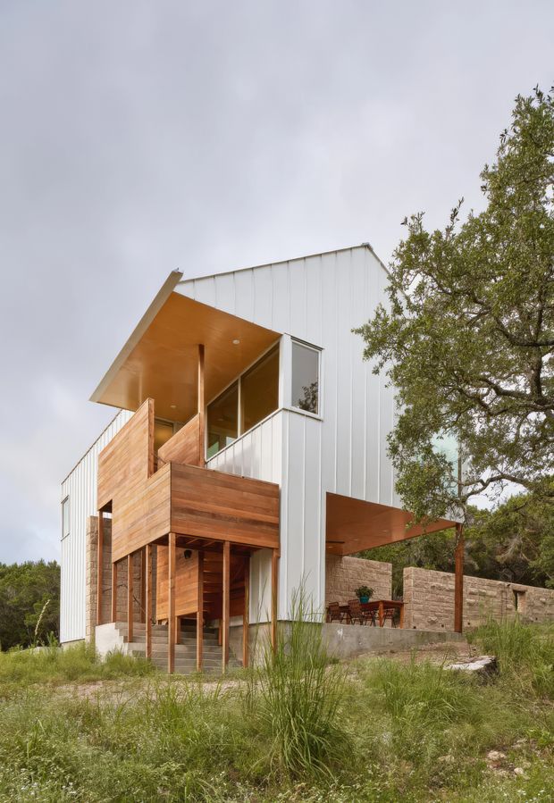 Exterior architectural photograph of a contemporary multi-level house on a natural, wooded and slightly sloped site. The building combines vertically ribbed white metal cladding with extensive warm timber cladding and soffits, projecting cantilevered roof forms, large glazed openings, exposed timber posts, balconies, and a sheltered outdoor dining area behind a low masonry wall.
