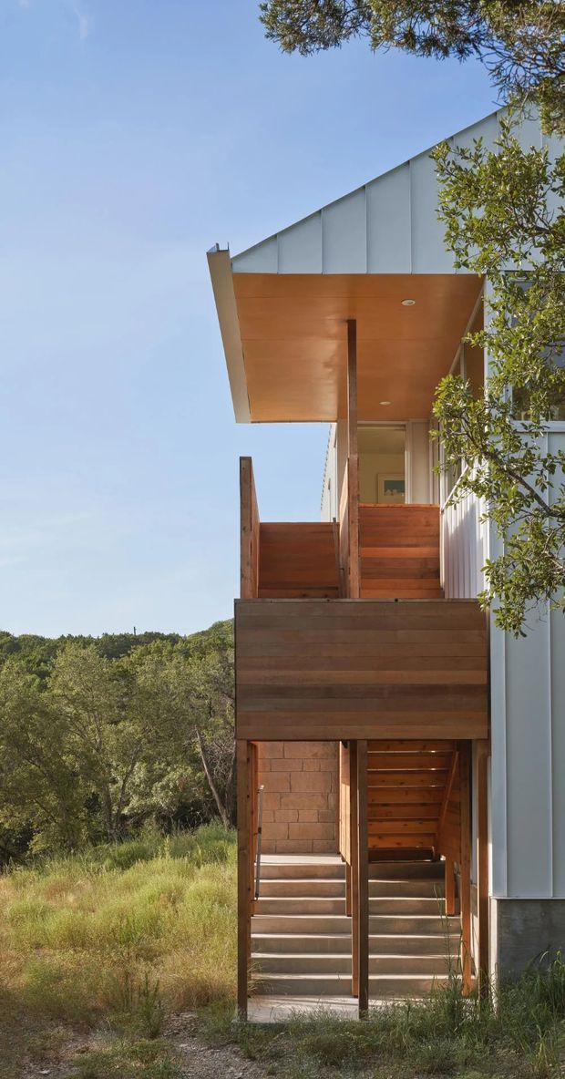 A vertically framed exterior view of a contemporary multi-level building set within a natural, bush-like landscape. The facade combines pale grey vertical metal cladding with extensive warm timber decking, soffits, screening and stair elements. A projecting upper canopy shelters a balcony, while an external stair and open timber structure connect the levels. Mature vegetation surrounds the building.