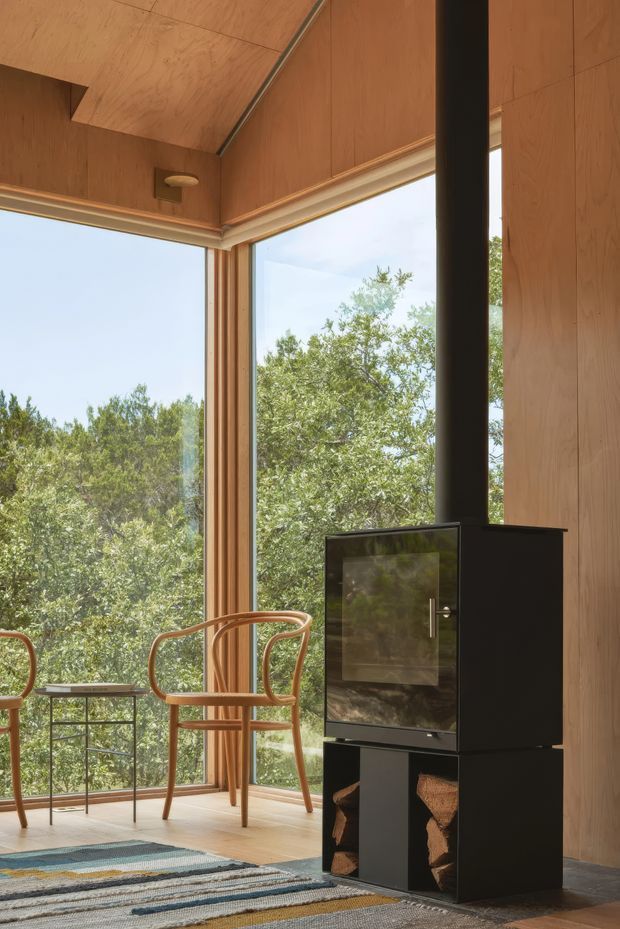 A warm, timber-lined interior corner with floor-to-ceiling corner glazing overlooking leafy vegetation. A black freestanding wood-burning stove with flue dominates the room, accompanied by two curved wooden armchairs, a small side table, and a patterned rug. The space has a calm, natural cabin-like atmosphere and abundant daylight.