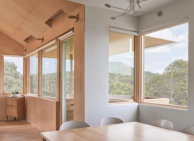 Bright contemporary dining area with a large light-timber dining table and chairs, extensive corner glazing, and warm timber wall and ceiling panelling. A compact timber storage cabinet sits near the windows, while a glazed door opens toward another room. The windows overlook a leafy, hilly landscape and bring abundant natural light into the space.