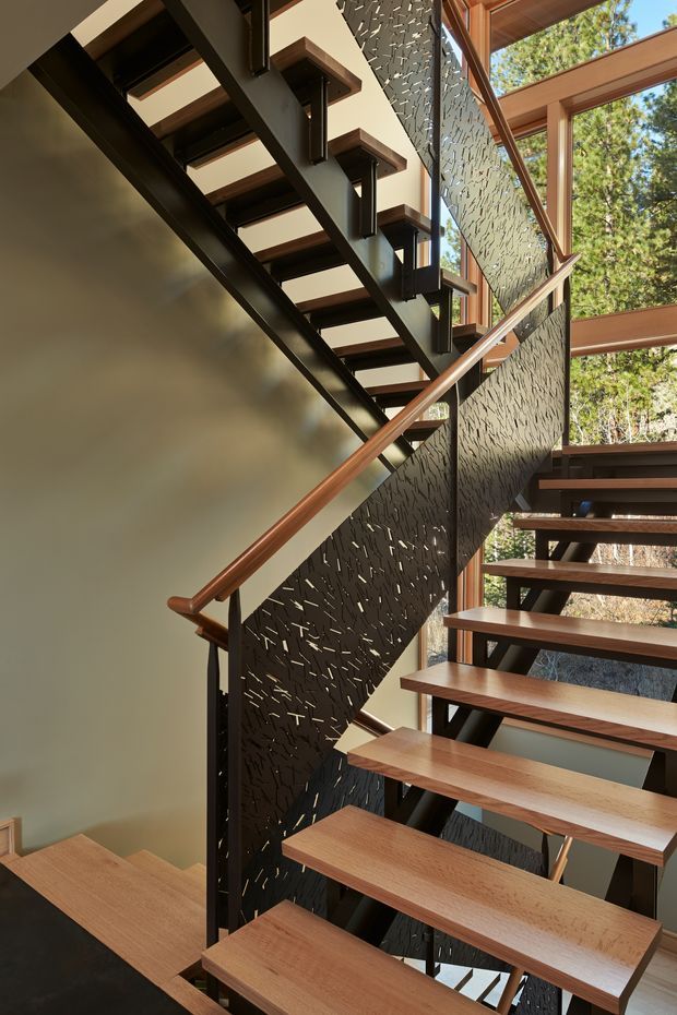 Interior view of a contemporary open-riser staircase with warm timber treads and handrail, dark painted steel stringers, and perforated patterned metal balustrade panels. The stair is arranged across multiple levels beside extensive timber-framed glazing, with views to mature trees outside. The composition highlights the sculptural stair structure, layered flights, and strong natural light.