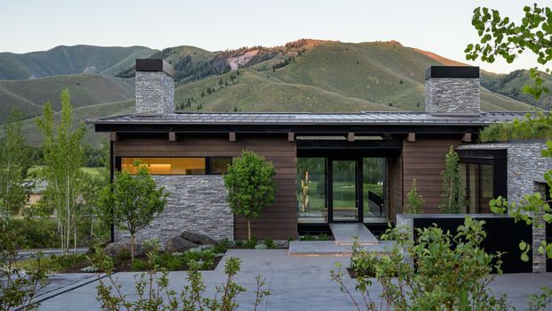 Low, single-storey contemporary residence set against a mountainous landscape. The facade combines dark horizontal timber cladding, stacked grey stone masonry, broad black-framed glazing, a shallow roof, and two prominent stone-clad chimney structures. A landscaped foreground with paved access and shrubs frames the home.