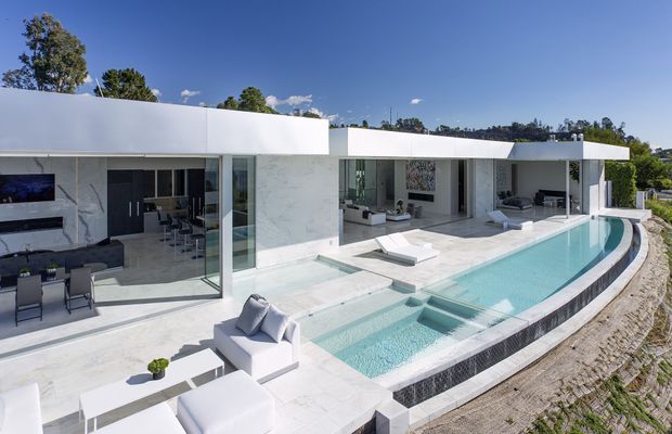 A luxurious modern single-storey residence with a crisp white flat-roofed facade, extensive floor-to-ceiling glazing, and a long curved-edge swimming pool. The bright white stone-clad outdoor terrace is furnished with streamlined white loungers and seating, while open-plan living and dining areas are visible through the glazing. The elevated setting looks across a tree-lined suburban landscape under a clear blue sky.