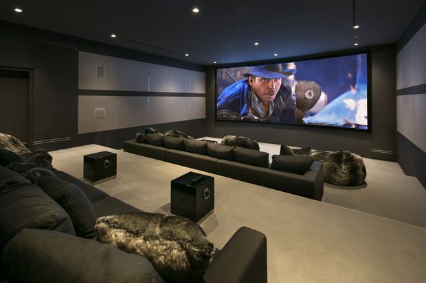 A dark, immersive home cinema with tiered or lounge-style seating arranged toward a large wall-mounted projection screen. The room features charcoal walls and ceiling, recessed downlights, light neutral flooring, acoustic wall panels, and multiple sofas with dark upholstery and textured throws.