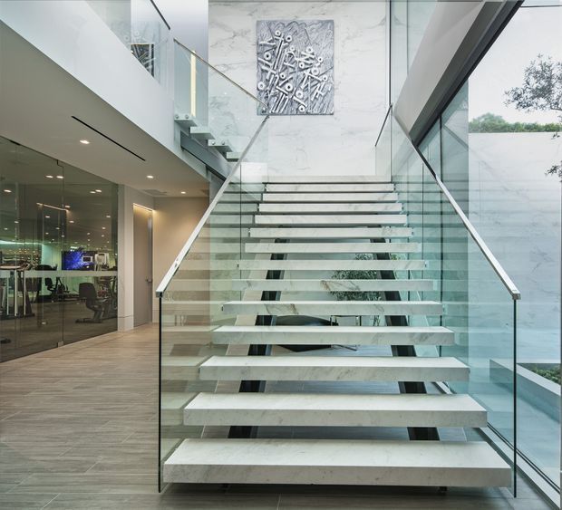 Contemporary interior featuring a dramatic floating staircase with pale stone treads, transparent glass balustrades, and a tall open stairwell. A glass-enclosed office or workspace is visible to the left, while marble-look wall surfaces, extensive glazing, and a large abstract artwork create a refined, light-filled setting.
