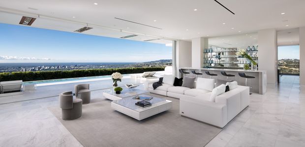 Bright, expansive modern living room with floor-to-ceiling glazing opening onto a terrace and pool, furnished with a large white sectional sofa, sculptural armchairs, and a low square coffee table. A sleek bar with glass shelving occupies the rear of the room, while the white marble-look flooring, pale neutral palette, recessed lighting, and panoramic coastal or urban outlook create a luxurious, airy atmosphere.