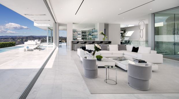Bright, spacious contemporary living room with a large white sectional sofa, sculptural lounge chairs, glass coffee tables, and a pale area rug. The room opens directly onto a white-paved terrace and pool area through extensive floor-to-ceiling glazing. A sleek bar or display joinery with glass shelving is visible at the rear, with a dramatic elevated landscape beyond.