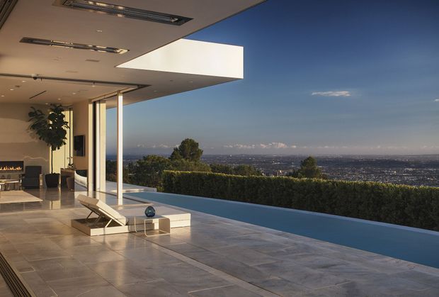 A luxurious contemporary indoor-outdoor living area opens onto a narrow infinity-edge pool or water feature, with expansive city and mountain views beyond. The space has large glazed openings, a projecting flat roof, pale stone-look flooring, minimalist outdoor loungers, and a warm interior with a linear fireplace and dining area.