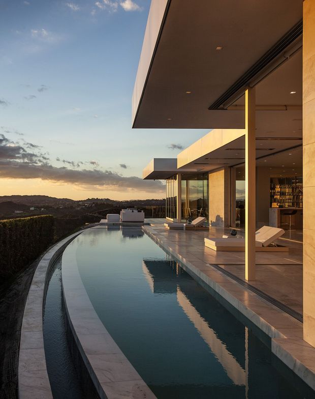 A contemporary luxury residence photographed at sunset, with a long infinity-edge swimming pool curving along the site beside a broad stone-paved terrace. The home features flat roof planes, extensive floor-to-ceiling glazing, slender structural supports, recessed exterior lighting, and warm light-toned stone cladding. An open-plan interior with a bar or kitchen area is visible through the glazing. The property overlooks a hilly landscape and distant urban settlement.