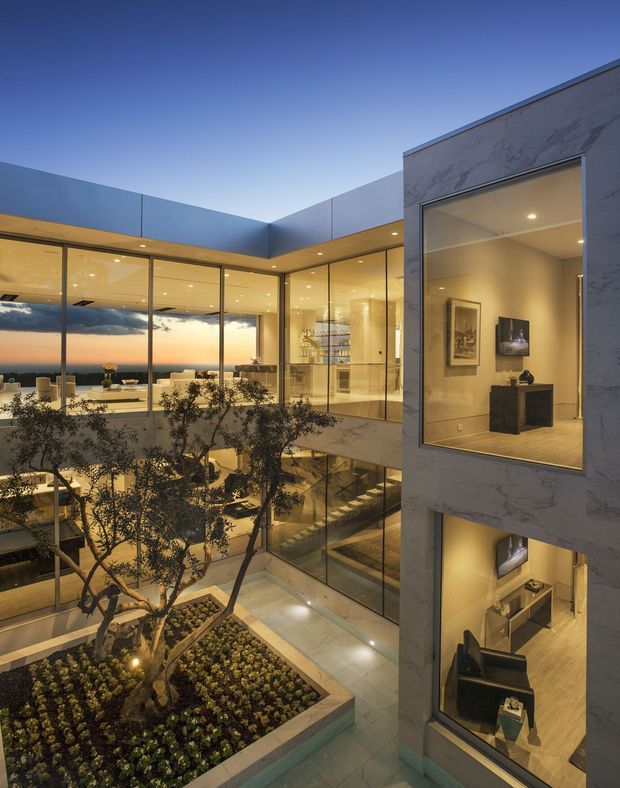 Twilight view into a sophisticated multi-storey contemporary residence organized around a landscaped internal courtyard. Extensive floor-to-ceiling glazing reveals warm-lit living and gallery-like spaces, while a pale stone-clad volume frames the right side. The courtyard includes raised planted beds and a small tree, with open balconies and stairs visible inside.