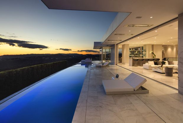 A luxurious contemporary indoor-outdoor living space opening onto a long infinity pool at sunset. The expansive terrace is finished in large pale stone tiles and furnished with modern white loungers, while floor-to-ceiling glazing connects it to a bright open-plan living area with a kitchen or bar, island bench, pendant lighting, and white lounge furniture. A broad cantilevered flat roof with recessed lighting shelters the terrace, with panoramic hills visible beyond the pool.