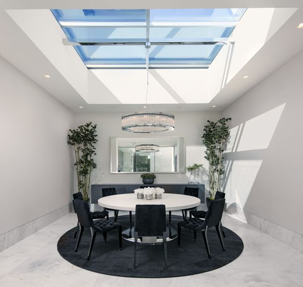 Bright, contemporary dining room with a round white dining table surrounded by dark upholstered chairs on a circular dark rug. The room has pale walls, a light stone or tiled floor, a large frameless skylight, recessed ceiling lights, a statement ring chandelier, tall indoor plants, and a large wall mirror reflecting the room.