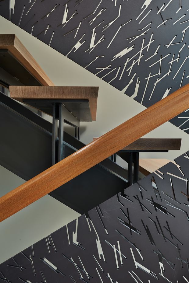 Looking upward through a sculptural multi-level stairway, with dark metal stair flights and balustrade panels cut with an irregular linear pattern. A prominent warm timber handrail crosses the composition, complemented by timber-edged landings and black structural supports. The image focuses on the stair circulation, material contrast, and decorative perforated screens.