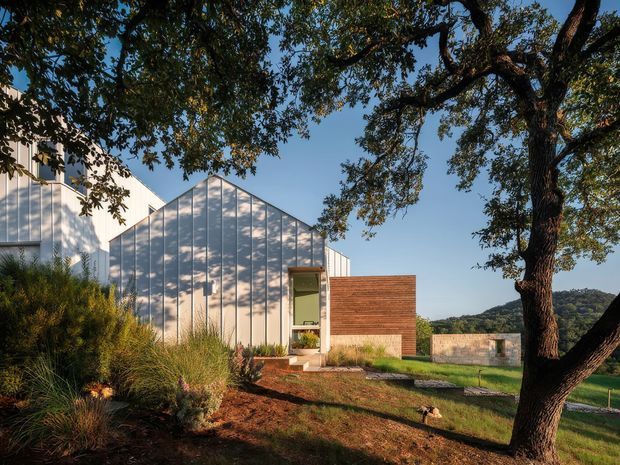A contemporary low-rise house set within a landscaped, sloping rural or semi-rural site. The building features a prominent gabled volume clad in light vertical panels, a projecting warm brick rectangular entry volume, and a simple glazed entrance. Native-style grasses and planting surround the house, with mature trees framing the view and hills visible in the distance.