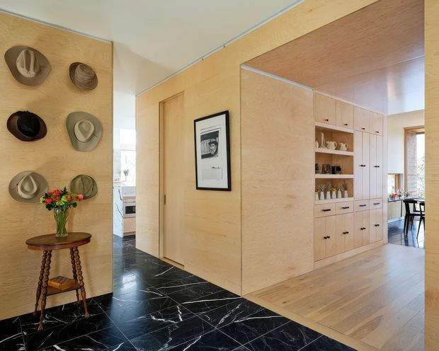 Interior view of a refined residential entry hall with pale timber wall panelling and built-in cabinetry. A dark marble-look tiled floor defines the hallway, transitioning to light timber flooring in the adjoining dining or living area. Decorative hats, a small timber console, framed artwork, shelving, and a glimpse of a dining table are visible. The composition highlights the transition between the entryway and the adjacent open room.