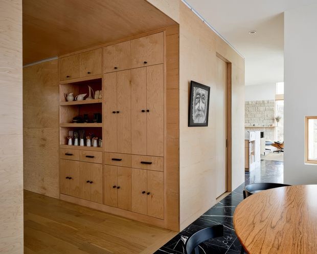 Warm contemporary interior showing a built-in timber storage and display wall with integrated cupboards and open shelving. The cabinetry sits beside a hallway leading toward a bright living area, with a round timber dining table and black chairs in the foreground. Pale timber wall and ceiling lining, timber flooring, dark stone-like floor tiles, and restrained black hardware create a refined natural palette.