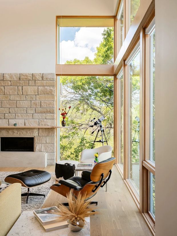 Bright, double-height living room with a full-height corner of timber-framed glazing overlooking leafy trees. A pale stone block fireplace with a simple mantel anchors the room, while a sculptural black leather lounge chair and ottoman, light upholstered chair, small table, and textured rug create a refined mid-century-inspired seating area. Natural timber flooring and warm wood window frames add warmth to the airy interior.