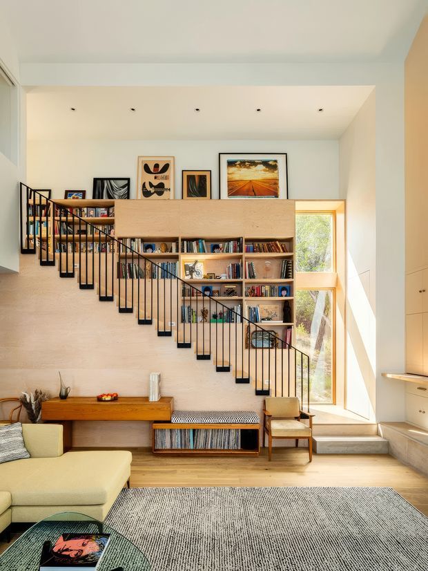 Bright, double-height living room with a sculptural stairway integrated into a full-height timber bookshelf. The room features pale textured walls, extensive built-in shelving, warm timber flooring and joinery, a large cream sectional sofa, woven rug, framed artwork, and tall glazing bringing in natural light.