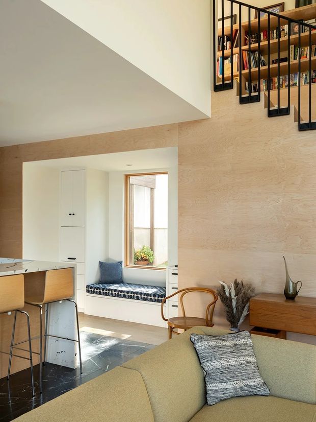 A warm, light-filled open-plan interior combining a living room, kitchen and dining area. Pale timber wall panelling continues around a stair with black metal balustrades and integrated open shelving. The room includes a curved neutral sofa, mid-century-style timber chair, timber sideboard, compact kitchen island with bar stools, a built-in window seat, and dark polished stone flooring.
