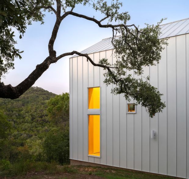 Exterior view of a compact contemporary building clad in pale vertical metal panels, with a simple pitched metal roof and narrow glowing amber windows. The structure sits within a heavily wooded, hilly landscape, partially framed by mature tree branches in the foreground.