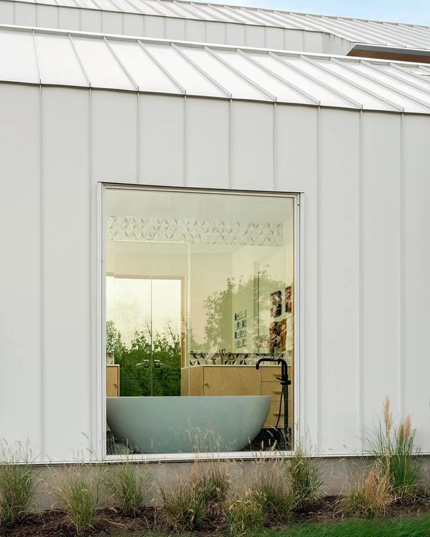 Exterior view of a contemporary small building clad in pale standing-seam metal, with a projecting or folded metal roof and a large glazed opening revealing a bathroom inside. The interior contains a freestanding bathtub, dark floor-mounted or wall-mounted tapware, timber vanity cabinetry, and patterned wall detailing. Ornamental grasses and low planting sit along the base of the facade.