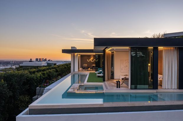 A modern flat-roofed residence viewed from an elevated terrace at sunset. The image is dominated by a rooftop infinity pool with crisp geometric edges, surrounded by pale paving and dark exterior wall bands. Floor-to-ceiling glazing opens onto a warmly lit interior with sheer curtains, while a distant urban skyline and dense treetops form the background.