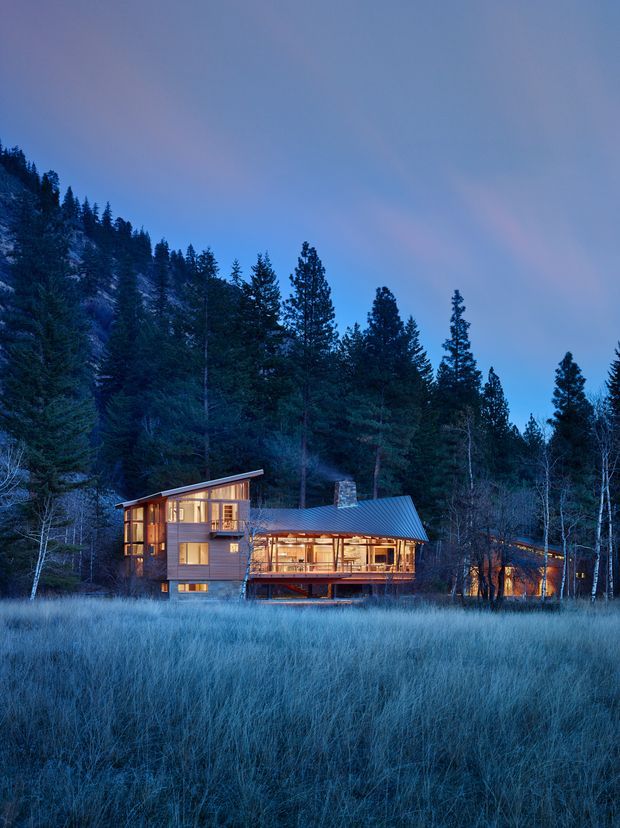 Twilight exterior photograph of a contemporary multi-level timber house set within a dense coniferous forest and meadow. The home features extensive warm-lit glazing, a projecting upper volume, a steeply sloped metal roof, exposed timber framing, and elevated deck-like living areas. The surrounding landscape is strongly wooded and alpine or mountain-like, with a cool blue dusk atmosphere contrasting against the warm interior lighting.