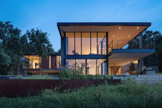 Twilight exterior view of a contemporary multi-level residence set among mature vegetation. The house features strong rectilinear volumes, a broad flat roof, extensive floor-to-ceiling glazing, dark structural framing, a projecting upper terrace with slender metal railings, and warm interior lighting visible through the windows. Landscaped planting and a low retaining wall occupy the foreground.