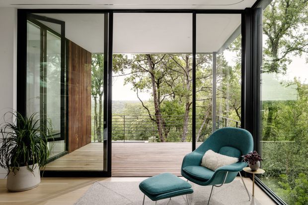 A contemporary interior opening onto a timber deck through expansive black-framed floor-to-ceiling glazing. The space features a teal upholstered lounge chair and ottoman, a pale rug, a potted plant, and a small side table, with leafy bushland views beyond the balcony. A vertical timber-clad wall adds warmth to the otherwise light, glass-filled room.