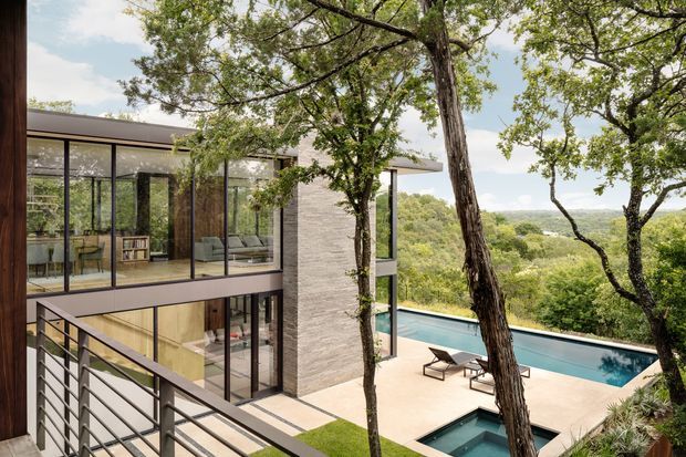 Exterior view of a contemporary multi-level home set among mature trees, with extensive floor-to-ceiling glazing, a pale stone-clad vertical wall, dark structural framing, and a terrace surrounding a long swimming pool. The image is taken from an elevated walkway or balcony and also reveals furnished interior living spaces through the glazing. A broad green, wooded landscape is visible beyond the property.