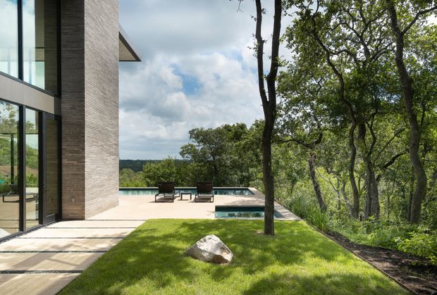 A contemporary house exterior beside a wooded, sloping landscape, viewed from a landscaped terrace. The image shows a tall glazed facade with dark framing and a textured brick or masonry wall, opening onto a pale paved pool terrace with lounge chairs and a rectangular swimming pool. A grassy lawn and mature trees occupy the foreground and right side, with distant wooded terrain beyond.