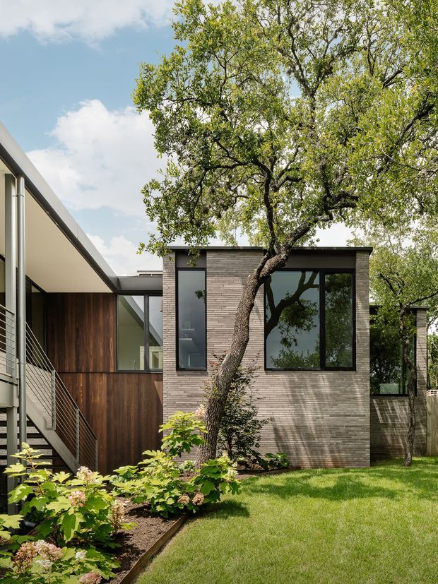 Exterior view of a contemporary residential addition or courtyard-facing house, featuring a low-pitched flat roof, pale horizontal brick masonry, expansive dark-framed glazing, and warm vertical timber cladding. An external metal stair and balcony occupy the left side, while mature trees, lawn, and landscaped planting surround the building.