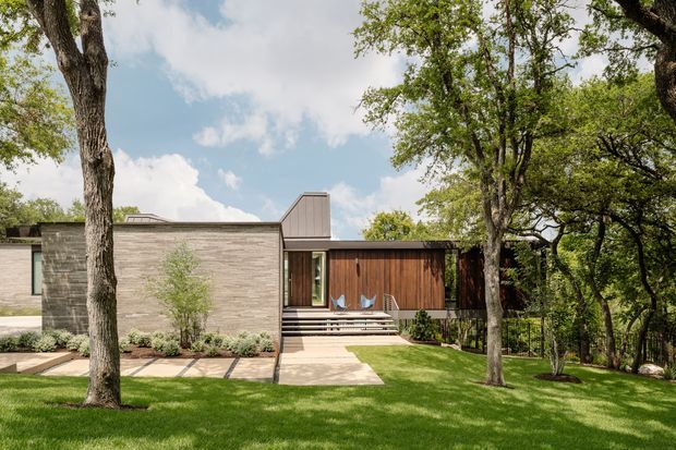 A low, single-storey modern residence set within a mature, leafy landscape. The facade combines pale horizontal masonry or stonework with warm vertical timber cladding, dark-framed glazing, and a small raised entry deck reached by broad steps. Large established trees, manicured lawn, planted borders, and concrete stepping slabs frame the house. The roofline is predominantly low and flat with a raised metal-clad chimney or roof element. The scene is photographed from the garden in bright daylight.