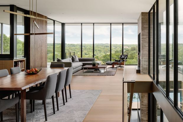 A refined open-plan living and dining area with expansive floor-to-ceiling glazing overlooking a leafy landscape. The room features a timber dining table, upholstered dining chairs, a large light-toned sectional sofa, a contemporary lounge arrangement, vertical timber wall panelling, pale timber flooring, and dark-framed glazing. A stair opening and internal glass balustrade are visible at the right.