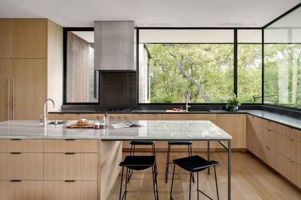 A contemporary kitchen with warm light-wood cabinetry, a large pale stone island with integrated sink and seating for two, black-framed floor-to-ceiling windows, and a dark tiled cooking backsplash beneath a stainless-steel range hood. The kitchen opens visually to leafy greenery outside and is furnished with two black counter stools.