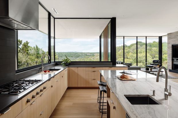 A spacious contemporary kitchen with extensive floor-to-ceiling black-framed glazing overlooking a leafy landscape. The room features warm timber cabinetry, dark stone or composite benchtops, a large pale veined stone island with integrated sink, a gas cooktop, stainless appliances, and streamlined black and timber detailing. Bar stools line the island, while a living area and fireplace are visible beyond.